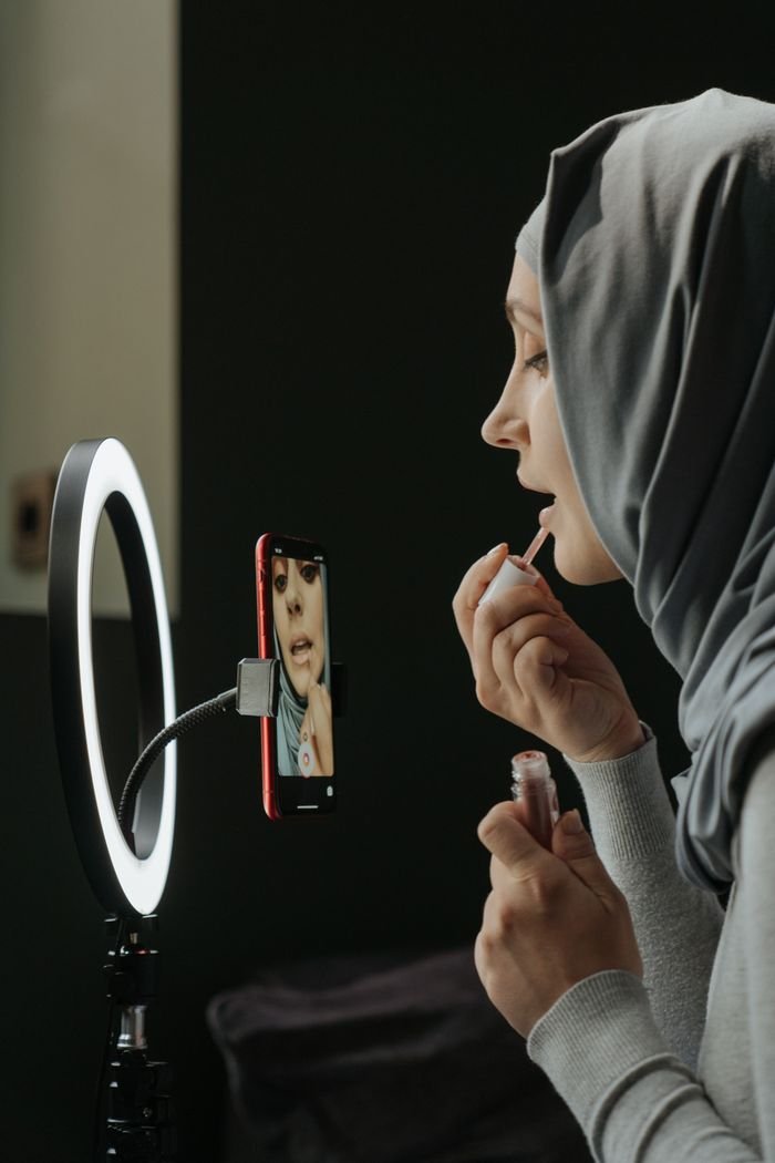 Woman in hijab applying lip gloss, using a smartphone and ring light for a beauty vlog.