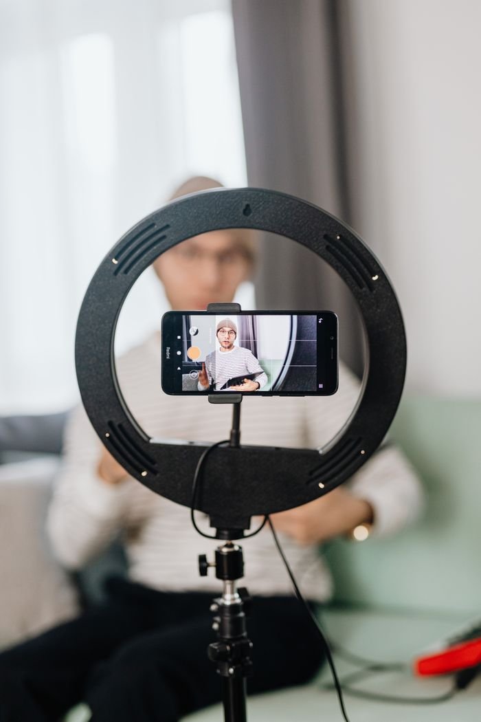 Seated man vlogging with a smartphone on a ring light tripod indoors.