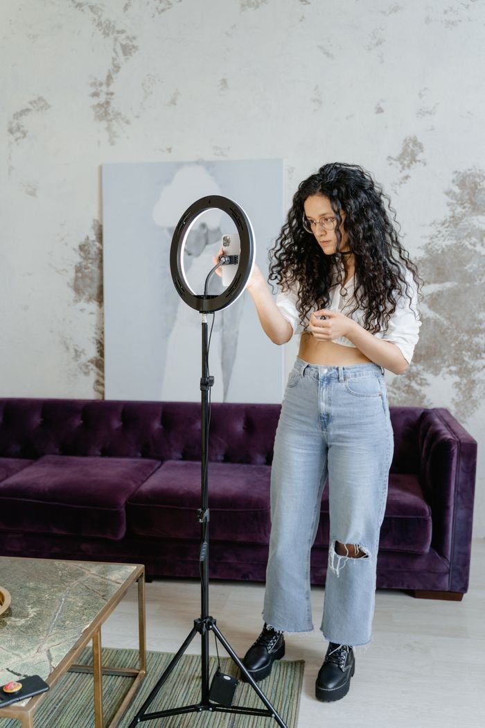 A young woman adjusts a ring light indoors, preparing for a social media video shoot.